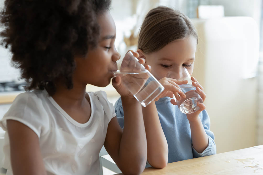 Two multiracial girls sit in kitchen feels thirsty drink water Two girls sitting in kitchen drinking purified water